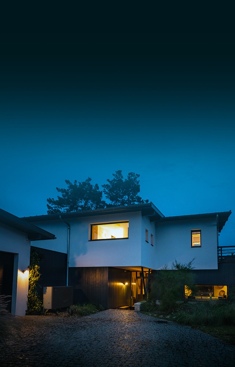 On a chilly winter evening, the overall exterior of a house with warm lights on. New black-colored LG Air to Water Heat Pump THERMA V is placed in front of the house. 	